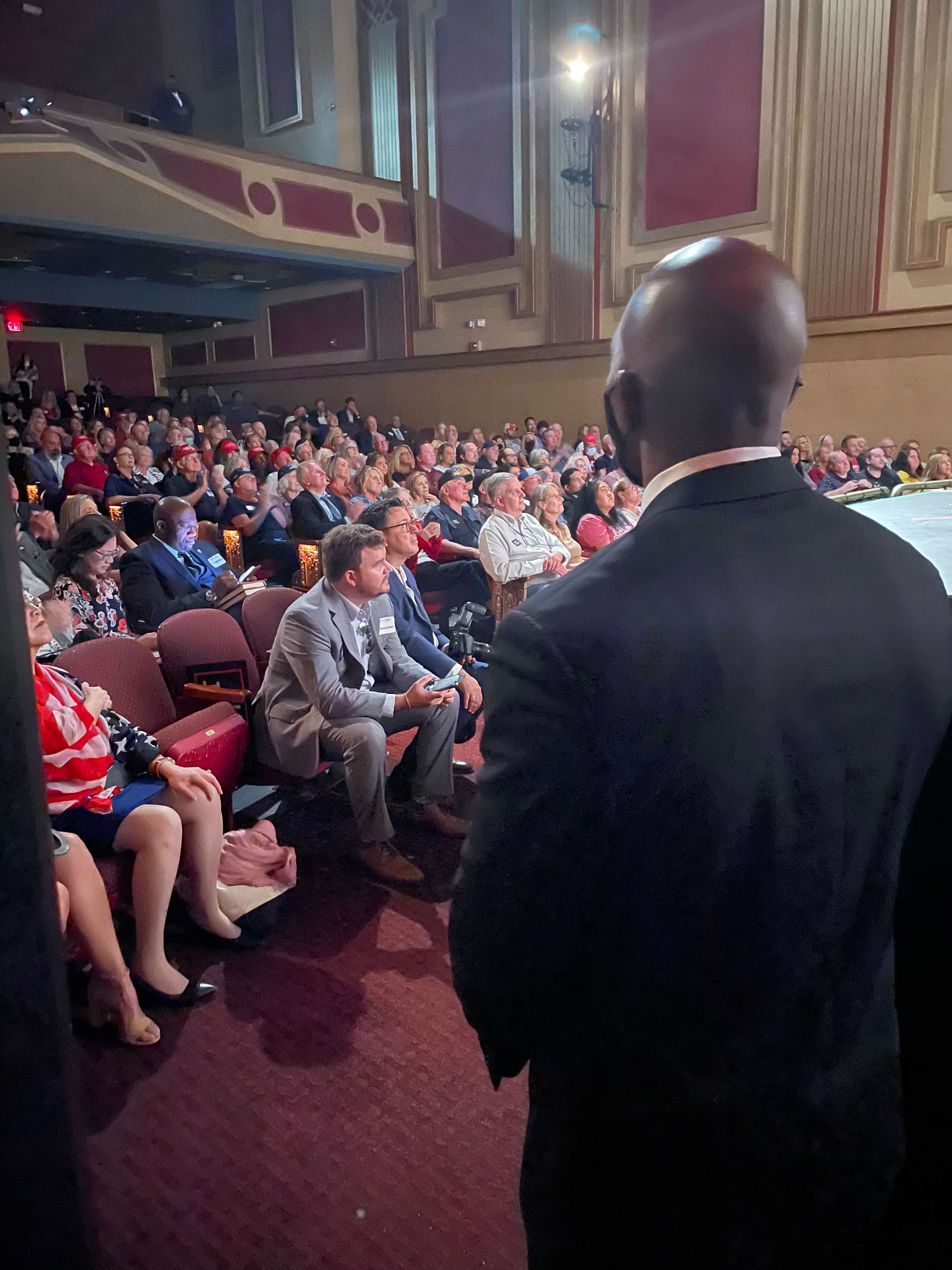 A suited Hawque protection professional overseeing a large political event from beside the stage